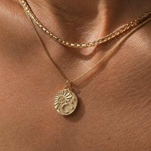 Puede incluir: Un conjunto de collares dorados en capas que consta de una cadena más gruesa y una cadena más fina con un colgante redondo. El colgante muestra un diseño en relieve de un sol, una luna creciente y pequeñas estrellas en su superficie circular.