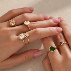 Könnte beinhalten: Eine Sammlung von sechs goldfarbenen Ringen an den Fingern. Die Ringe haben verschiedene Designs, darunter runde und quadratisch geschliffene klare Steine, einen quadratischen dunkelgrünen Stein und einen kleinen dunkelblauen Stein.