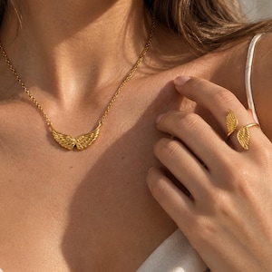 May include: A gold-toned necklace featuring a central pendant shaped like a pair of spread angel wings on a delicate chain, paired with a matching open-style ring that also displays two small, detailed angel wing motifs on each end of the band.
