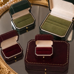Puede incluir: Una colección de cuatro joyeros de terciopelo en verde oscuro y burdeos intenso. Las cajas presentan formas octogonales, forros interiores de color crema y cierres de metal dorado. Los insertos de terciopelo sujetan los anillos de forma segura.