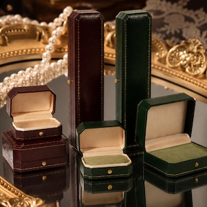 Puede incluir: Una colección de joyeros rectangulares en material similar al cuero de color burdeos intenso y verde oscuro. El conjunto incluye estuches verticales altos y cajas más anchas con tapas abatibles, cierres de metal dorado e interiores de terciopelo.