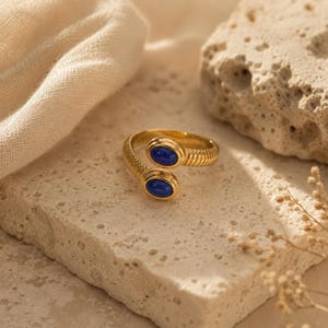 Puede incluir: Un anillo abierto de tono dorado con una banda texturizada y dos piedras ovaladas de color azul intenso en cada extremo. El anillo presenta un diseño abierto donde los extremos no se tocan, con un acabado metálico pulido y cabujones azules lisos.