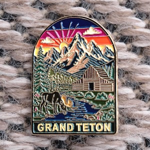 May include: An arched enamel pin featuring a colorful mountain landscape with a sunrise, a log cabin, a moose near a stream, and evergreen trees. The sky transitions from yellow to orange and pink. The text GRAND TETON is printed in gold at the bottom.