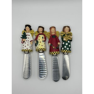 May include: A set of four decorative spreaders featuring angel figurines as handles. Each angel wears a unique outfit with a checkered hem and holds a holiday-themed item, including a green tree, a candle, a heart, and a gold star.