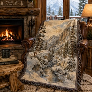 Könnte beinhalten: Eine gewebte Wohndecke mit einer detaillierten Winterlandschaft, die schneebedeckte Felsen, immergrüne Bäume und einen fließenden Bach zeigt. Die Decke hat einen Fransenrand und stellt ein Gebirge in gedeckten Farben dar.