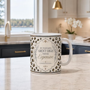Puede incluir: Una taza de cerámica blanca con un estampado de leopardo en negro y dorado. El centro muestra un marco decorativo con el texto: MY STANDARDS AREN'T HIGH THEY'RE Expensive, seguido del texto: THE GRACE COLLECTION y un pequeño logotipo.