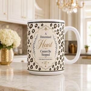 May include: A white ceramic mug featuring a black and gold leopard print pattern. The center displays a decorative frame with the text A Determined Heart Cannot Be Stopped, The Grace Collection, and the initials LL in gold and black lettering.