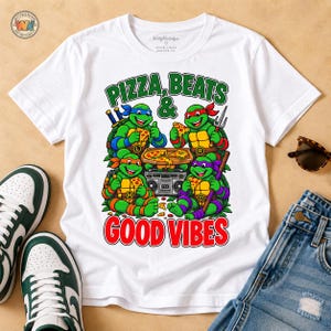 May include: A white short-sleeve t-shirt featuring a graphic of four green turtles with colored masks eating pizza around a boombox. The text reads PIZZA, BEATS & GOOD VIBES. The turtles are labeled with letters L, R, M, and D on their chest plates.