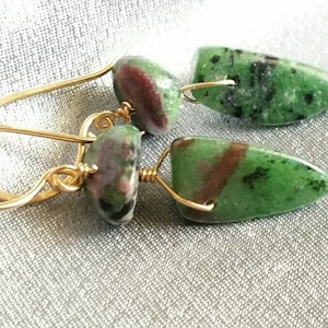 May include: A pair of gold earrings with green, pink, and black gemstone drops. The earrings are made with wire wrapped around the stones.