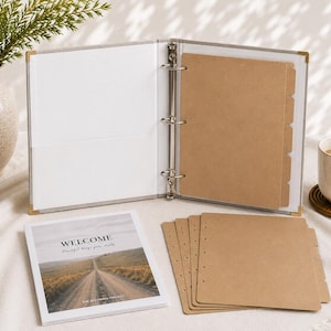 May include: A light gray three-ring binder with gold-tone corner protectors, containing brown kraft paper dividers. A square booklet with a landscape photo and the text "WELCOME Beautiful things grow slowly THE BEGINNING PROJECT" sits beside loose brown paper sheets.