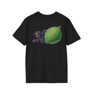 May include: A black short-sleeve t-shirt featuring a graphic print on the chest. The design depicts a small, dark purple creature with glowing pink eyes and a yellow flower on its head, positioned next to a large, bright green lime fruit.