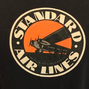 Może przedstawiać: Czarny T-shirt z białym okrągłym logo przedstawiającym zabytkowy samolot przed pomarańczowym słońcem. Logo brzmi "Standard Air Lines".