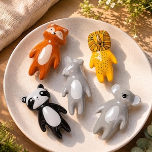 May include: Five ceramic animal-shaped chopstick rests arranged on a speckled cream plate. The figures include an orange fox, a yellow lion with a black mane, a grey donkey, a black and white panda, and a light grey koala, each with a white belly.