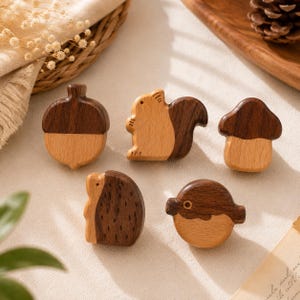 May include: Five wooden cabinet knobs featuring two-tone light and dark wood finishes. The shapes include an acorn, a squirrel, a mushroom, a hedgehog, and a round bird. Each piece is crafted with smooth, polished surfaces and distinct wood grain.