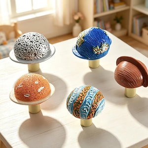 Op de afbeelding: Een set van vijf decoratieve knoppen in de vorm van hemellichamen, waaronder een zilveren maan met kraters, een blauw-groene aarde, een oranje planeet met textuur, een gestreepte blauw-beige planeet en een bruine planeet, elk op een cilindrische voet.