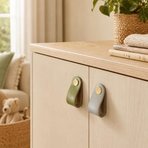 May include: Two cabinet pulls made of folded leather straps, one in olive green and one in light gray, each secured with a circular brass-toned metal fastener. The pulls are mounted on a light-colored wooden cabinet door with a smooth finish.