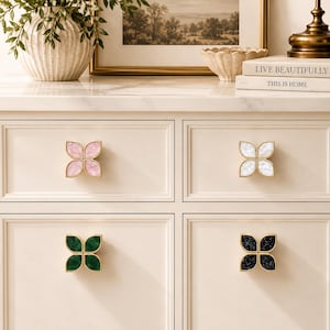 May include: Four flower-shaped cabinet knobs with gold-toned metal frames. Each knob features four petal-like inserts in distinct colors: one with light pink, one with white, one with dark green, and one with black, all with a subtle speckled texture.