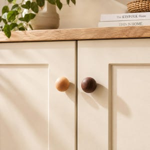 Peut inclure: Deux boutons de placard sphériques fixés sur des portes de placard couleur crème. Un bouton est en bois clair et l'autre en bois foncé. Les boutons sont ronds et lisses, positionnés au centre des panneaux avant des placards.