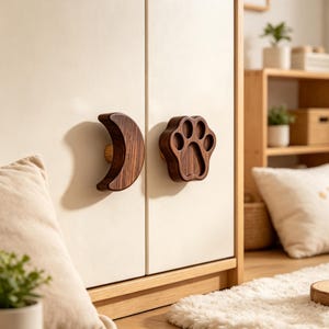May include: Two wooden cabinet knobs attached to a white cabinet door. One knob is shaped like a crescent moon, and the other is shaped like a paw print. Both knobs feature a dark brown wood grain finish and are mounted on small, light-colored wooden bases.
