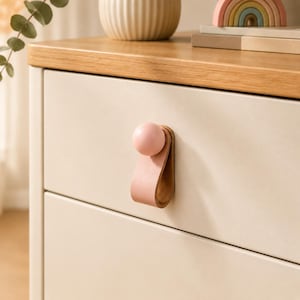 Puede incluir: Un tirador de cajón que presenta un lazo de cuero rosa suave asegurado por un pomo de madera redondo a juego. El herraje está montado en un frente de cajón liso y de color claro, proporcionando un mango funcional con un diseño redondeado.