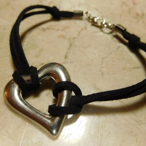 Può includere: Un braccialetto in corda di camoscio nero con un ciondolo a forma di cuore d'argento. Il cuore è aperto e la corda è infilata attraverso di esso.