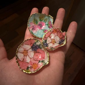 K&ouml;nnte beinhalten: Drei dekorative Muschelschalen mit goldfarbenen R&auml;ndern. Jede Muschel ist mit einem floralen Decoupage-Muster aus rosa, wei&szlig;en und blauen Blumen auf hellem Grund verziert. Die Muscheln sind konkav geformt und weisen unterschiedliche Gr&ouml;&szlig;en auf.