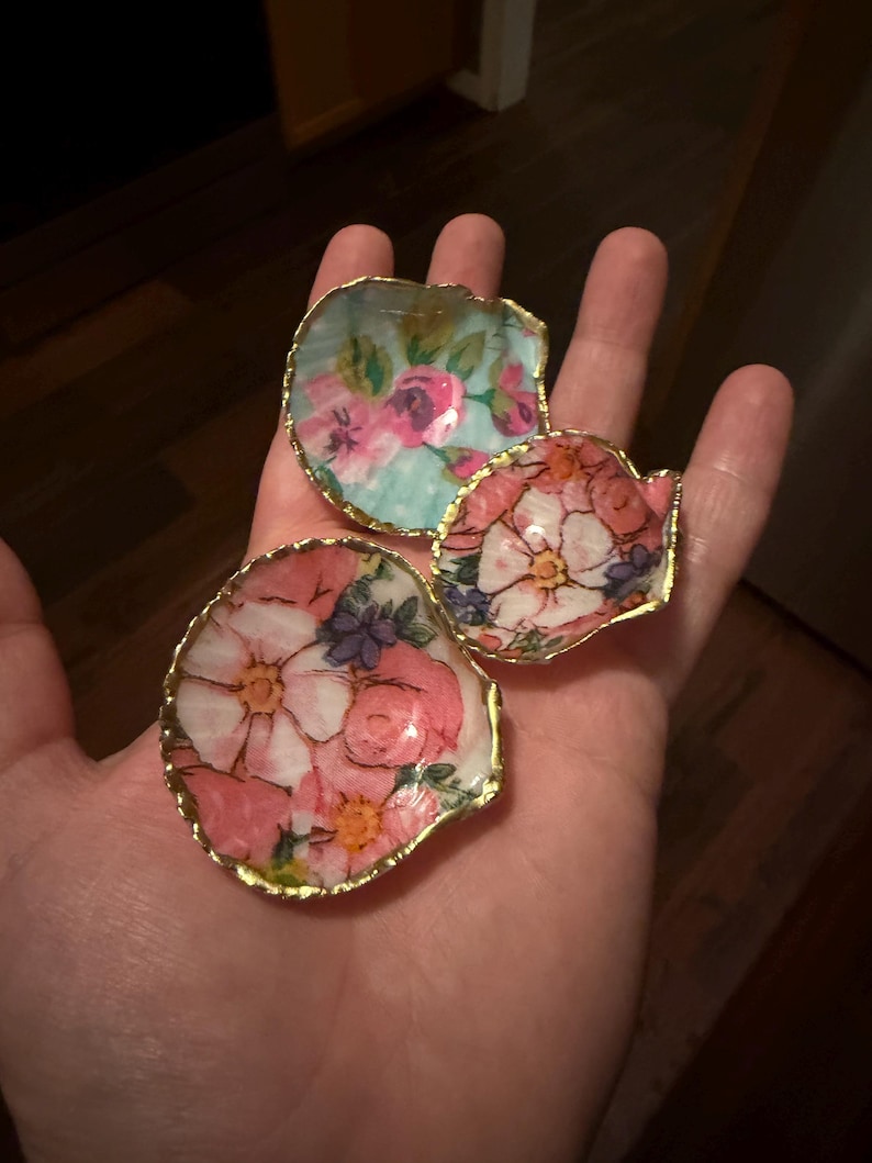 K&ouml;nnte beinhalten: Drei dekorative Muschelschalen mit goldfarbenen R&auml;ndern. Jede Muschel ist mit einem floralen Decoupage-Muster aus rosa, wei&szlig;en und blauen Blumen auf hellem Grund verziert. Die Muscheln sind konkav geformt und weisen unterschiedliche Gr&ouml;&szlig;en auf.