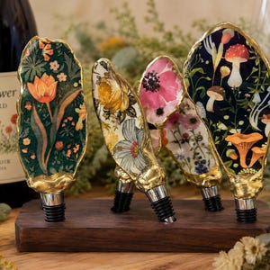 May include: Four decorative bottle stoppers with gold-painted edges, each featuring unique botanical and mushroom illustrations on the front. The stoppers are displayed upright in a rectangular dark wood stand with four circular cutouts.
