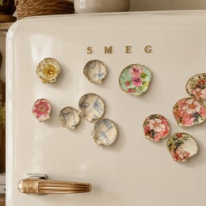 K&ouml;nnte beinhalten: Eine Sammlung dekorativer Muschelmagnete an einem cremefarbenen SMEG-K&uuml;hlschrank. Jede Muschel zeigt ein einzigartiges florales oder blaues Schleifenmuster in Decoupage-Technik mit goldfarbenen, gewellten R&auml;ndern, die in einer Gruppe angeordnet sind.