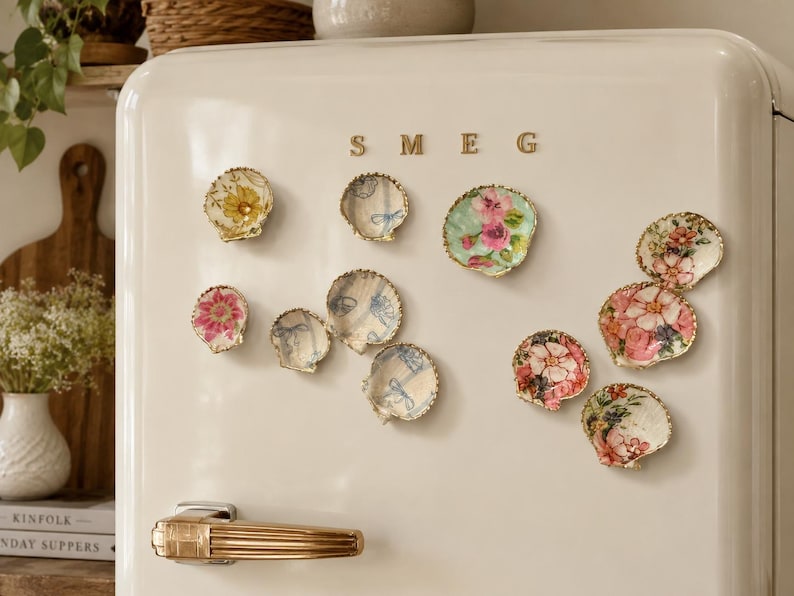 K&ouml;nnte beinhalten: Eine Sammlung dekorativer Muschelmagnete an einem cremefarbenen SMEG-K&uuml;hlschrank. Jede Muschel zeigt ein einzigartiges florales oder blaues Schleifenmuster in Decoupage-Technik mit goldfarbenen, gewellten R&auml;ndern, die in einer Gruppe angeordnet sind.