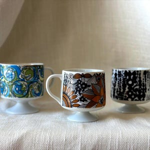 Puede incluir: Tres tazas de cerámica con pedestal y patrones distintos. La taza de la izquierda presenta diseños de remolinos azules y verdes. La taza central muestra motivos florales naranjas y negros. La taza de la derecha tiene un patrón geométrico en blanco y negro.