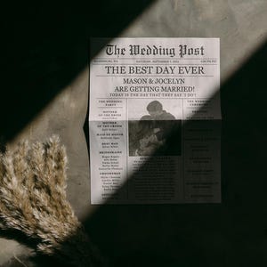 May include: A printed newspaper-style wedding program titled The Wedding Post, featuring the text Mason & Jocelyn Are Getting Married, a central photograph of a couple, and lists of the wedding party members, accompanied by a dried pampas grass stem.