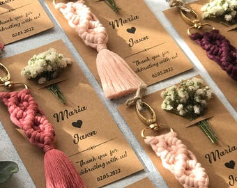 May include: A collection of macrame keychains in shades of pink and deep purple, each attached to a gold-tone lobster clasp. The keychains are mounted on rectangular kraft paper cards printed with the names Maria and Jason and the date 02.05.2022.