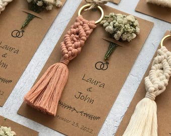 May include: Macrame keychains with gold-tone metal rings attached to rectangular kraft paper cards. Each card features a small dried flower bouquet, the names Laura & John, a wedding ring icon, and the date September 23, 2022, in black ink.