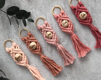 May include: Five macrame keychains in varying shades of pink and red, each featuring a gold-toned metal ring, a central wooden bead engraved with a name, and a knotted cord design ending in a tassel. The names include Naomi, Valeria, Dana, Emily, and Kaila.