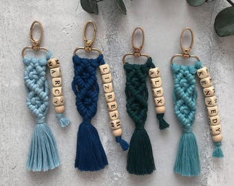 May include: Four macrame keychains in shades of blue, each featuring a gold-tone lobster clasp, a knotted cord design, and a strand of light-colored wooden beads with black letters spelling the names Marcy, Lizbeth, Alex, and Kennedy.
