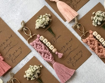 May include: Five macrame keychains in shades of pink and cream, each attached to a brown card with dried baby's breath flowers. The cards feature the text 'Will you be my Bridesmaid?' and individual names including Martha, Dana, and Valeria.