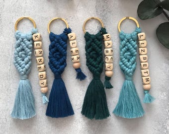 May include: Four macrame keychains in shades of light blue, navy blue, dark green, and teal. Each keychain features a gold metal ring, a knotted cord design, a matching tassel, and a vertical strand of wooden beads with the names KENNEDY, ALEX, MARCY, and LIZBETH.