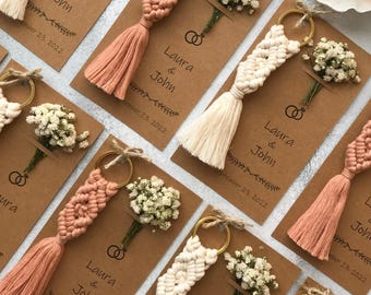 May include: Macrame keychains in cream and dusty rose colors attached to brown cardstock tags. Each tag features the printed text Laura & John, a date, a graphic of two interlocking rings, and a small dried baby's breath flower bouquet.