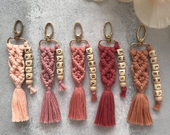 May include: Five macrame keychains in varying shades of pink, ranging from light peach to deep terracotta. Each keychain features a gold-tone lobster clasp, a knotted cord design, a tassel, and a wooden bead strand spelling out names: KARLA, DANA, MARTHA, EMILY, and VALERIA.