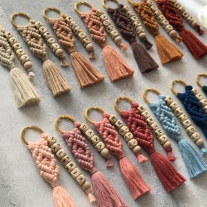 May include: A collection of macrame keychains in various colors, each featuring a gold-toned metal ring, a knotted cord design, and wooden beads with printed letters spelling names such as Karla, Maria, Katie, Naomi, Gabby, Audrey, Laura, Valeria, Dana, Emily, Martha, Kennedy, and Alex.