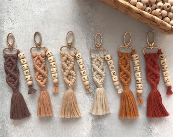 May include: Six macrame keychains in varying shades of brown, tan, and rust, each featuring a gold-tone metal clasp, a knotted cord design, a matching tassel, and a vertical string of wooden letter beads spelling names like Laura, Katie, and Maria.