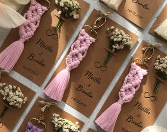 May include: Macrame keychains in shades of pink and purple attached to rectangular kraft paper cards. Each card features the printed text Martha & Brendan 02.09.2021, an infinity symbol, and a small dried flower bouquet tied with twine.