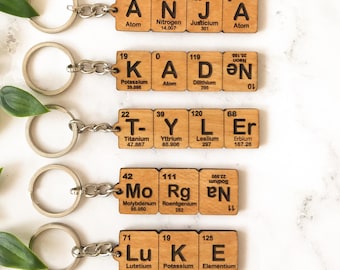 May include: Five wooden keychains featuring square tiles with black engraved letters and numbers, mimicking periodic table elements. Each keychain displays a name: ANJA, KADEN, TYLER, MORGAN, and LUKE, attached to a silver metal split ring.