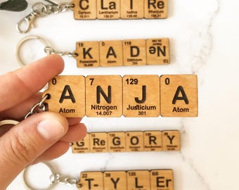May include: A collection of wooden keychains featuring names spelled out using square tiles designed to resemble periodic table elements. Each tile displays a letter, a chemical symbol, an atomic number, and a name, attached to a metal ring.