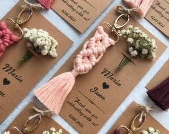 May include: Macrame keychains in shades of pink and dark purple, each featuring a gold-tone lobster clasp and a small bouquet of dried white baby's breath flowers. The keychains are attached to brown cardstock tags printed with Maria, Jason, and 02.05.2022.