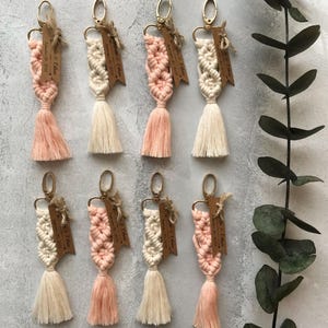 May include: Eight macrame keychains featuring knotted cotton cord in alternating shades of cream and soft peach. Each keychain includes a gold-toned metal lobster clasp and a small, rectangular brown wooden tag engraved with text.
