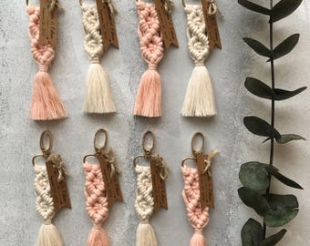 May include: Eight macrame keychains featuring knotted cotton cord in alternating shades of cream and soft peach. Each keychain includes a gold-toned metal lobster clasp and a small, rectangular brown wooden tag engraved with text.