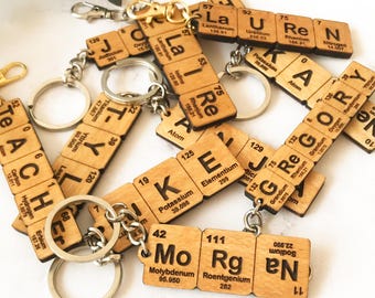 May include: A collection of custom keychains made from light-colored wood squares, each printed with chemical element symbols and atomic numbers from the periodic table to spell out names like Lauren, Gregory, and Morgan, attached to metal rings.