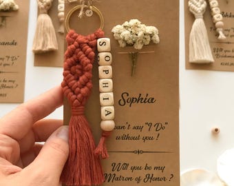 May include: A terracotta macrame keychain with a gold ring and wooden beads spelling SOPHIA, attached to a brown card featuring dried white flowers and the printed text: I can't say I Do without you! Will you be my Matron of Honor? 02.05.2025.